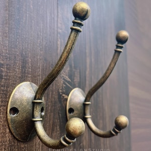 Op de afbeelding: Twee antieke messing wandhaken met een gebogen ontwerp en kogelvormige uiteinden. De haken zijn gemonteerd op ovale achterplaten en staan tegen een donkere houten achtergrond. De haken zijn ongeveer 15 cm hoog.