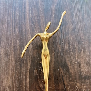 May include: A golden metal sculpture of a stylized figure with arms outstretched. The figure has a slender body and is posed in a graceful, dancing position. The sculpture is set against a dark wood background. The text "ECHODESIGNSTUDIO" is at the bottom.