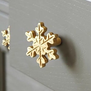 Può includere: Primo piano di un cassetto con finitura grigio chiaro e tiretti a forma di fiocco di neve dorati. Il cassetto è aperto e l'interno è in legno. Il cassetto è etichettato "Kitchen & Beyond" con il testo "handcrafted by Lundins Kök & Interiör" sotto.