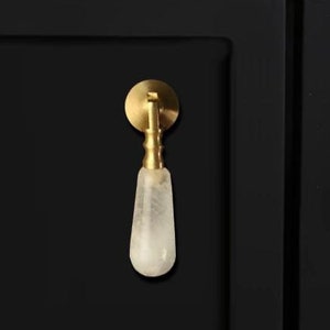 May include: A white crystal cabinet knob with a gold metal base and a round back plate. The knob is mounted on a black cabinet door.