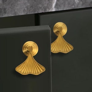 Może przedstawiać: Dwa złote uchwyty do szafki w kształcie liścia miłorzębu z wypukłym, fakturowanym wzorem. Uchwyty są zamontowane na ciemnoszarych drzwiach szafki.