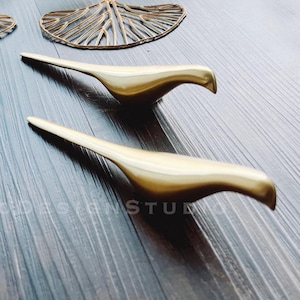 May include: Two gold metal bird sculptures on a dark wood surface. The birds are stylized and have a sleek, modern design.