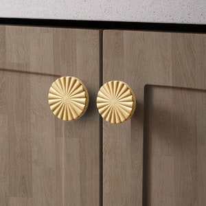 Puede incluir: Dos pomos redondos de color dorado para armario con un diseño de sol. Los pomos están montados en una puerta de armario de madera.