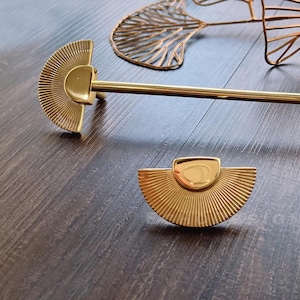 Könnte beinhalten: Goldene Metall-Schrankknöpfe mit einem fächerförmigen Design. Die Beschläge haben ein dekoratives, geometrisches Muster.