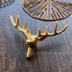 Könnte beinhalten: Goldfarbener Metall-Wandhaken in Form eines Hirschkopfes mit Geweih. Der Haken befindet sich auf einem dunklen Holzgrund.