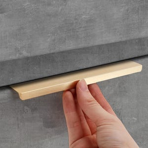 May include: A gold-colored cabinet pull handle is shown against a gray cabinet. The handle is rectangular with a flat front and a slightly curved edge. The handle is being held by a person's hand.