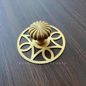 Peut inclure: Un bouton de meuble doré avec un dessus arrondi et cannelé et une plaque arrière circulaire décorative. La plaque arrière présente un motif ajouré de formes interconnectées. Le bouton et la plaque arrière sont en métal. Le texte "ECHO DESIGN STUDIO" est visible.