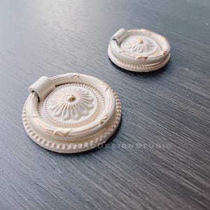 Peut inclure: Deux poignées de tiroir en céramique blanche avec une bordure dorée et un motif floral en relief. Les poignées ont une forme de bague pour une prise en main facile.