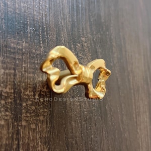 May include: A gold-colored ring in the shape of a bow. The ring has a textured, flowing design. The ring is on a dark brown wooden surface. The text "ECHODESIGNSTUDIO" is visible.