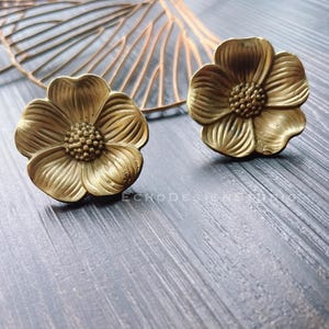 May include: Two brass-colored flower-shaped cabinet knobs with detailed petal designs. The knobs are on a gray wooden surface, with a decorative leaf in the background. The knobs are likely for furniture.