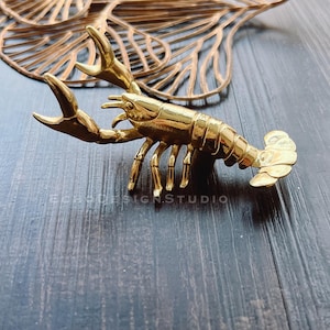May include: A gold-colored lobster figurine with detailed claws, legs, and tail. The lobster is positioned on a dark wooden surface, with a decorative gold leaf design in the background. The text "ECHO DESIGN STUDIO" is visible.