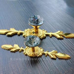 以下が含まれることがあります： 透明なファセットクリスタルノブが付いた、2つの金色のキャビネットプル。プルは、花のモチーフを持つ装飾的なデザインです。「ECHODESIGNSTUDIO」というテキストが表面に表示されています。