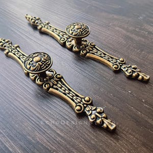 Può includere: Due tiretti per ante di armadio in tinta oro, decorati con intricati motivi floreali. Ogni tiretto presenta un pomello rotondo con un motivo floreale in rilievo.