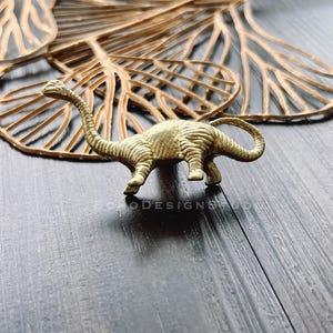 May include: A gold-coloured dinosaur figurine with a long neck and tail, standing on a dark wooden surface with a decorative, light brown, leaf-shaped background.