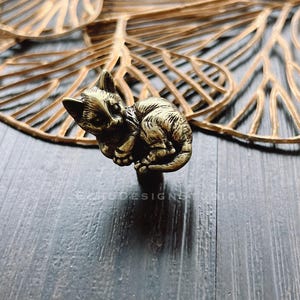 May include: A gold-toned metal cat figurine door knob. The cat is curled up in a sleeping position. The knob is on a dark wood surface.