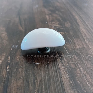 Op de afbeelding: Witte keramische kastknop met een afgeronde vorm en een zwarte metalen basis.