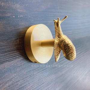 Peut inclure: Crochet mural en forme d'escargot doré avec une base ronde. Le crochet est en métal et a une finition texturée.