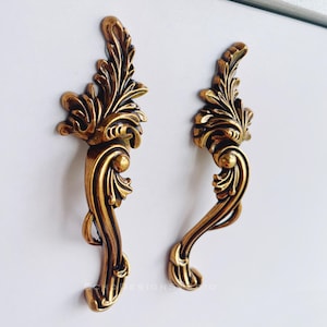 May include: Two ornate, antique-style cabinet handles. The handles are made of gold-colored metal and feature a detailed, leaf-like design. The handles are mounted on a white surface, showcasing their intricate details.
