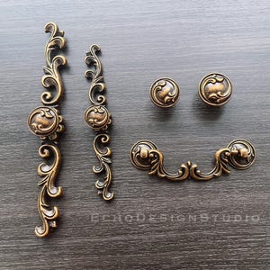 Op de afbeelding: Een assortiment antieke messing kastbeslagen, waaronder twee lange grepen, twee knoppen en een handvat. De hardware heeft sierlijke krullen en een warme, verouderde afwerking, perfect om een vintage touch aan meubels toe te voegen.