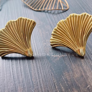 May include: A pair of gold-tone earrings shaped like a fan. The earrings are made of metal and have a shiny finish.