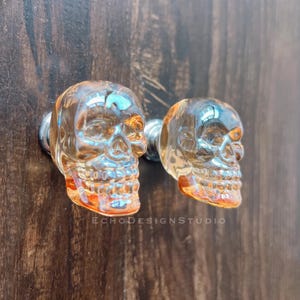 May include: Two clear glass skull cabinet knobs with a metallic silver base. The knobs are mounted on a dark brown wooden surface. The text "ECHODESIGNSTUDIO" is visible on the surface.