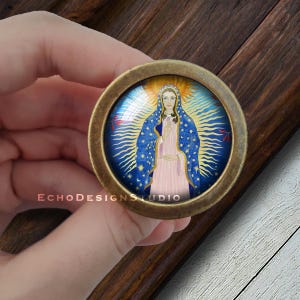 Może przedstawiać: Okrągły, mosiężny gałka z wkładką szklaną przedstawiającą kolorowy obraz Matki Boskiej. Obraz przedstawia ją w niebieskiej i złotej szacie z różową sukienką, stojącą na niebieskim i złotym tle z gwiazdami.