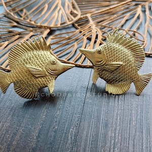 Op de afbeelding: Twee gouden metalen vissen met gedetailleerde schubben en vinnen. De vissen kijken in tegengestelde richtingen en liggen op een donker houten oppervlak.