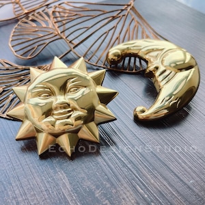 Peut inclure: Deux ornements en métal doré, un soleil et une lune croissante, sur une surface en bois. Le soleil a un visage avec un sourire et les yeux fermés. La lune est une simple forme de croissant.
