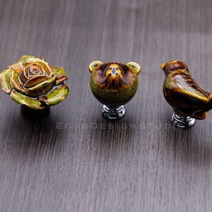Puede incluir: Tres tiradores decorativos para armarios: una rosa, un oso y un pájaro. El tirador de la rosa es un diseño detallado de varias capas con tonos verdes y marrones. El tirador del oso tiene un cuerpo redondo con orejas de oso. El tirador del pájaro es marrón con detalles amarillos.
