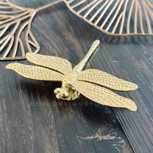 Puede incluir: Un pomo de puerta de libélula dorado con acabado texturizado. La libélula es un elemento de diseño popular para la decoración del hogar.