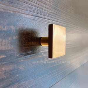 Peut inclure: Un bouton de porte carré en or avec une base arrondie. Le bouton est monté sur une surface en bois foncé.