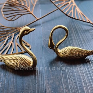 May include: Two gold swan-shaped decorative objects on a dark wood surface. The swans are facing opposite directions.