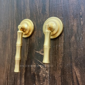 Può includere: Due pomelli dorati per mobili con un design ispirato al bambù. Ogni pomello presenta una piastra posteriore circolare e una maniglia a forma di steli di bambù. I pomelli sono montati su una superficie di legno marrone scuro. Il testo "ECHODESIGN STUDIO" è visibile.