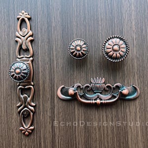 May include: Three antique-style cabinet knobs with ornate designs. The knobs are made of metal and have a copper finish. One knob is a pull handle with a floral design. The other two knobs are round with a floral design.