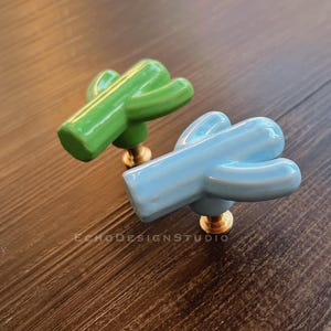 May include: Two ceramic cactus cabinet knobs. One is light green, the other is light blue. Both have a brass base. The knobs are on a dark wood surface. The text "ECHODESIGNSTUDIO" is visible.