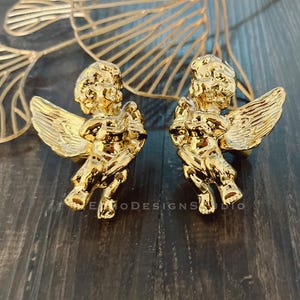 Könnte beinhalten: Zwei goldfarbene Schrankknöpfe in Form von Engeln mit detaillierten Flügeln. Sie befinden sich auf einer dunklen Holzoberfläche, mit einem dekorativen goldenen Blattdesign im Hintergrund. Geeignet für Möbel oder Dekoration.