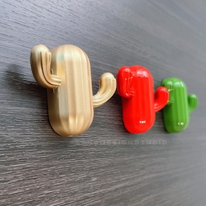 Può includere: Tre ganci a forma di cactus in oro, rosso e verde. I ganci sono montati su una superficie in legno scuro.
