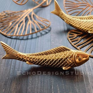 May include: Two gold-colored fish sculptures with detailed scales. The fish are on a wooden surface with a gold-colored leaf design.