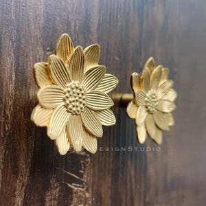 Può includere: Due pomelli per mobili dorati a forma di girasole. I pomelli sono in metallo e presentano dettagliati disegni di petali e centro. I pomelli sono montati su una superficie di legno marrone scuro. Il testo "DESIGNSTUDIO" è visibile.