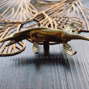 Puede incluir: Una figurita de dinosaurio de metal de color dorado con un hocico largo y una superficie texturizada. El dinosaurio está en posición de carrera con la cola levantada.