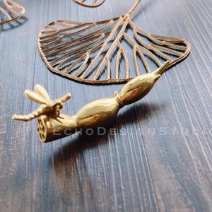 May include: Gold-toned metal dragonfly sculpture with a detailed design. The dragonfly is perched on a branch with a textured surface. The sculpture is on a dark wood background.