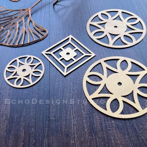 May include: A collection of laser-cut brass metal shapes, including circular, oval, and square geometric designs. A copper-colored leaf shape is also present. The shapes are arranged on a dark blue wooden surface. The text "ECHODESIGNSTU" is visible.