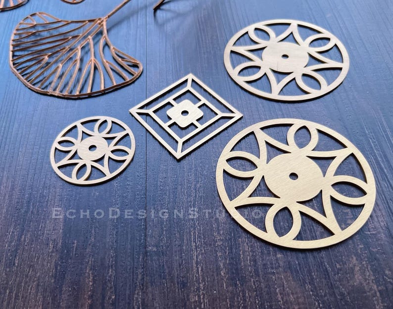May include: A collection of laser-cut brass metal shapes, including circular, oval, and square geometric designs. A copper-colored leaf shape is also present. The shapes are arranged on a dark blue wooden surface. The text "ECHODESIGNSTU" is visible.
