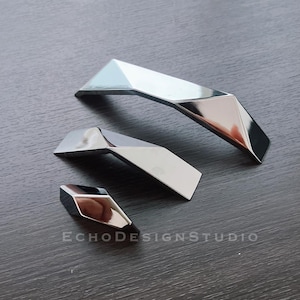 Op de afbeelding: Drie zilverkleurige kastdeurgrepen in verschillende maten en vormen. De grepen hebben een geometrisch ontwerp.