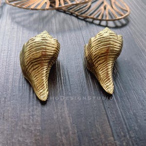 May include: Two gold-coloured seashell-shaped decorative objects. The shells have a textured surface with detailed ridges. The objects are placed on a dark wooden surface, with a blurred decorative element in the background.