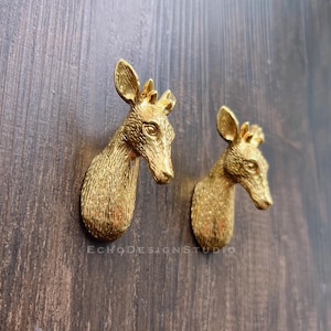 Könnte beinhalten: Zwei goldfarbene Schrankknöpfe in Form von Hirschköpfen. Die Hirschköpfe sind nach rechts gerichtet und haben eine detaillierte, strukturierte Oberfläche.