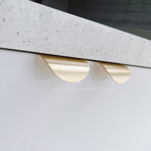 May include: Close-up of two brushed brass cabinet pulls with a curved, petal-like design. The pulls are mounted on a white cabinet door, with a speckled gray countertop above. The text "ECHODESIGNSTUDIO" is visible.