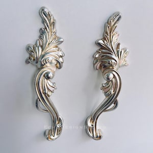 May include: A pair of silver-coloured, decorative door handles. Each handle features a leaf-like design at the top, with a curved body and a decorative ball. The handles are set against a white background.