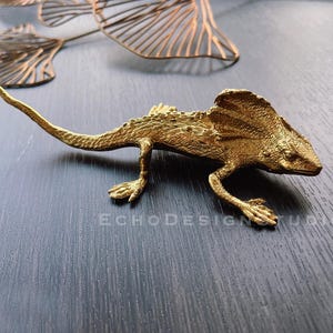 Puede incluir: Una figurita de lagarto de color dorado con una gola alrededor del cuello. La figurita está en posición de caminar con la cola extendida detrás de ella.