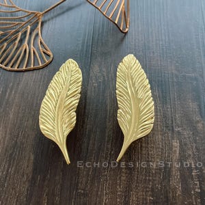 Könnte beinhalten: Zwei goldfarbene, federförmige Schubladengriffe aus Metall. Die Griffe befinden sich auf einer dunkelbraunen Holzoberfläche.